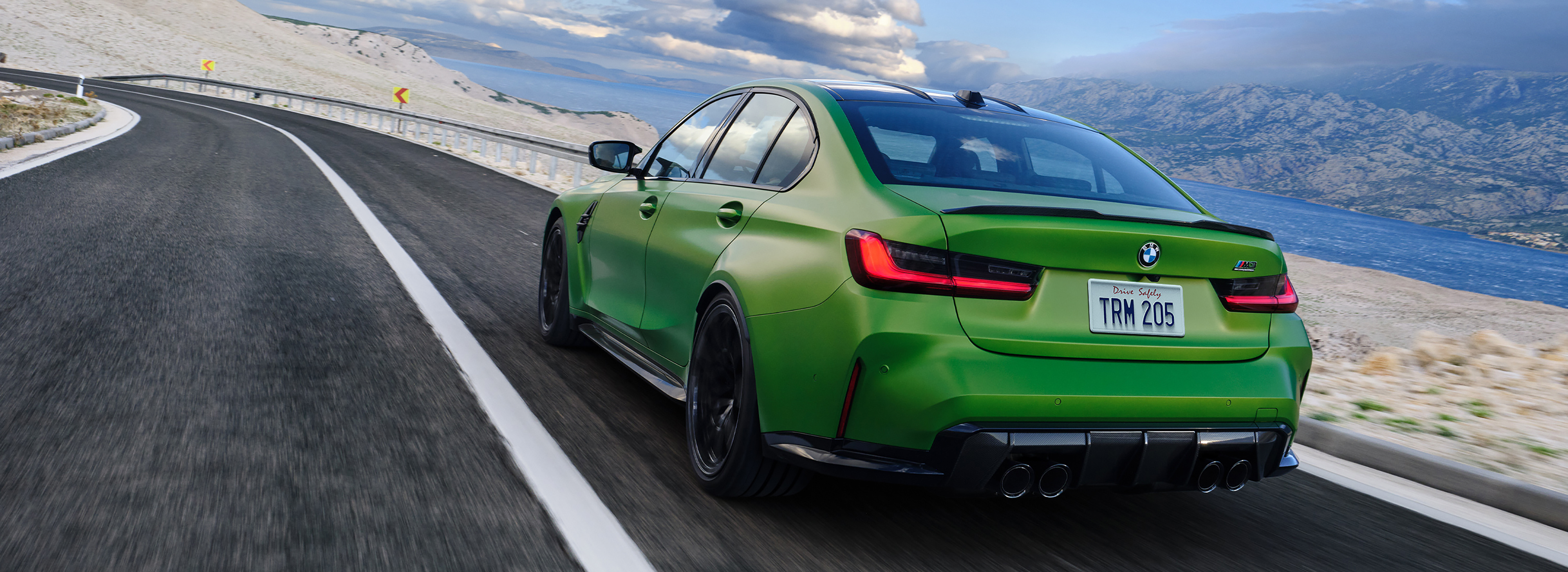 BMW M3 sports sedan driving along a coastal mountain road, showcasing its green exterior, performance design, and dynamic handling.