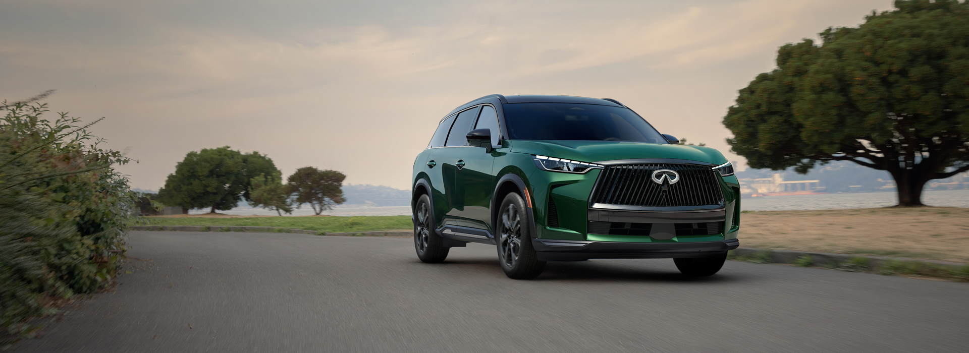 Front view of a green Infiniti SUV driving along a scenic road, with trees and water in the background.