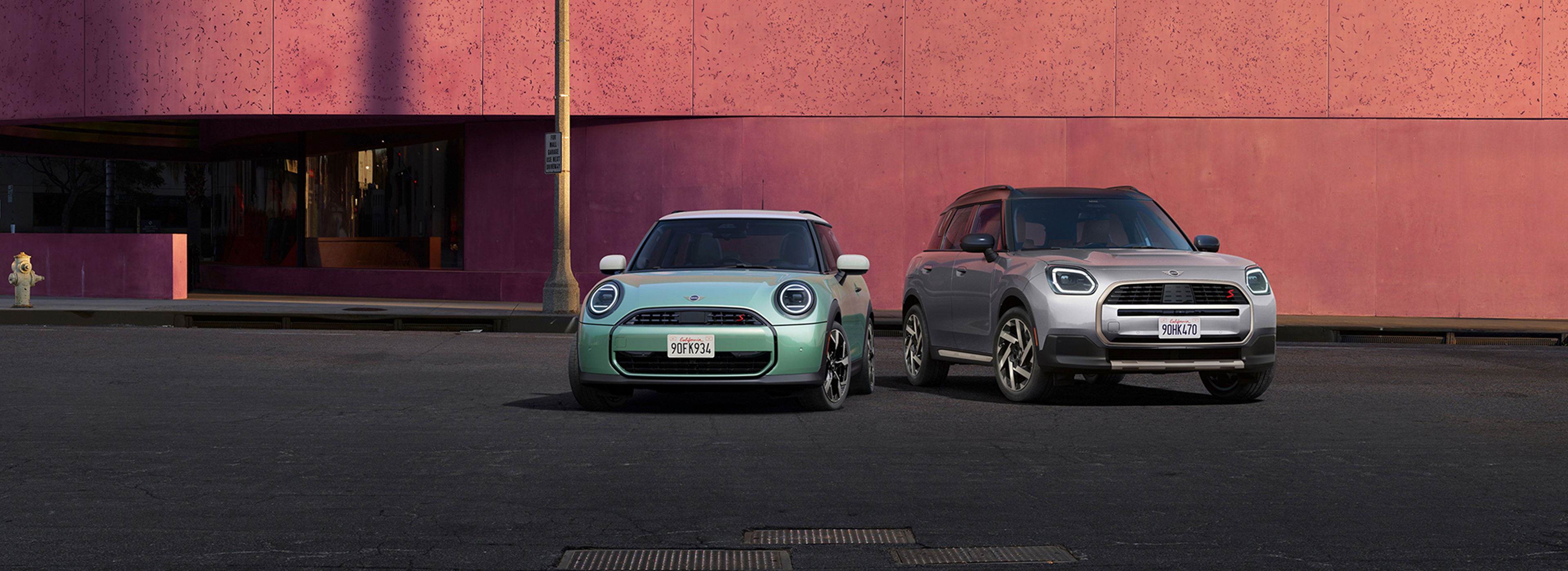 Two MINI vehicles—a mint-green MINI Cooper and a silver MINI Countryman—parked side by side in front of a modern pink building.