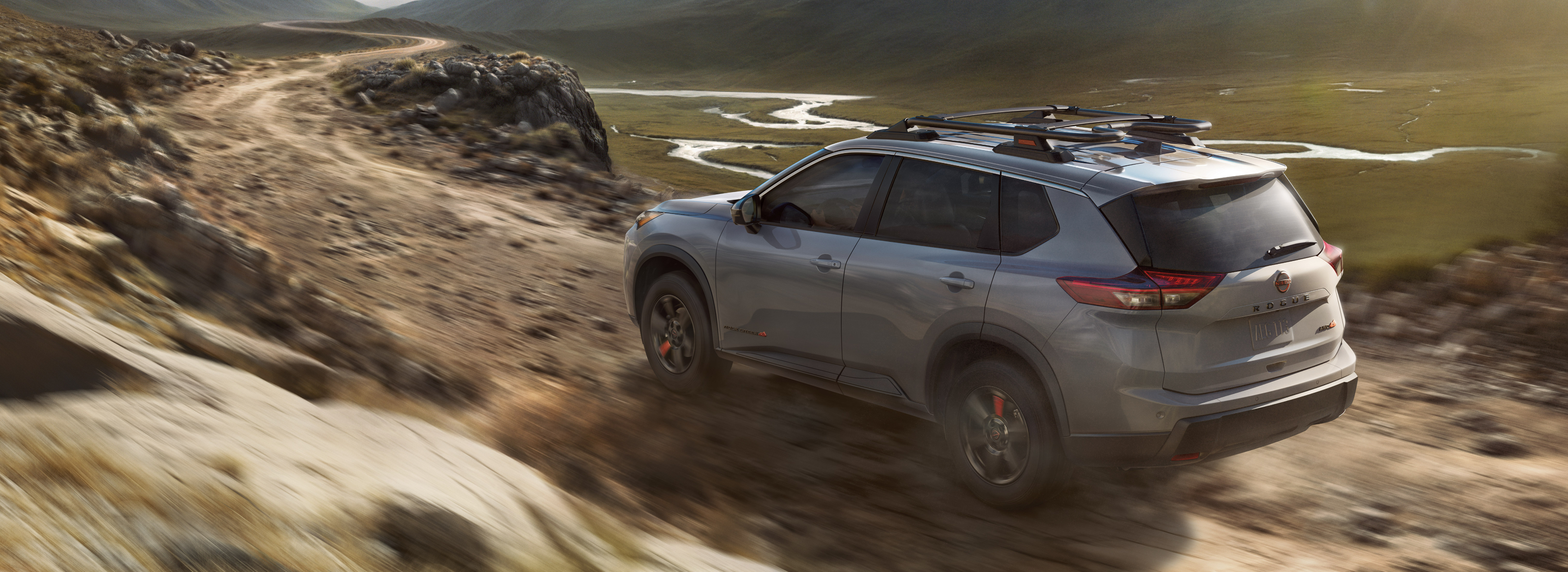 Nissan Rogue driving off-road through rugged mountain terrain, kicking up dust.