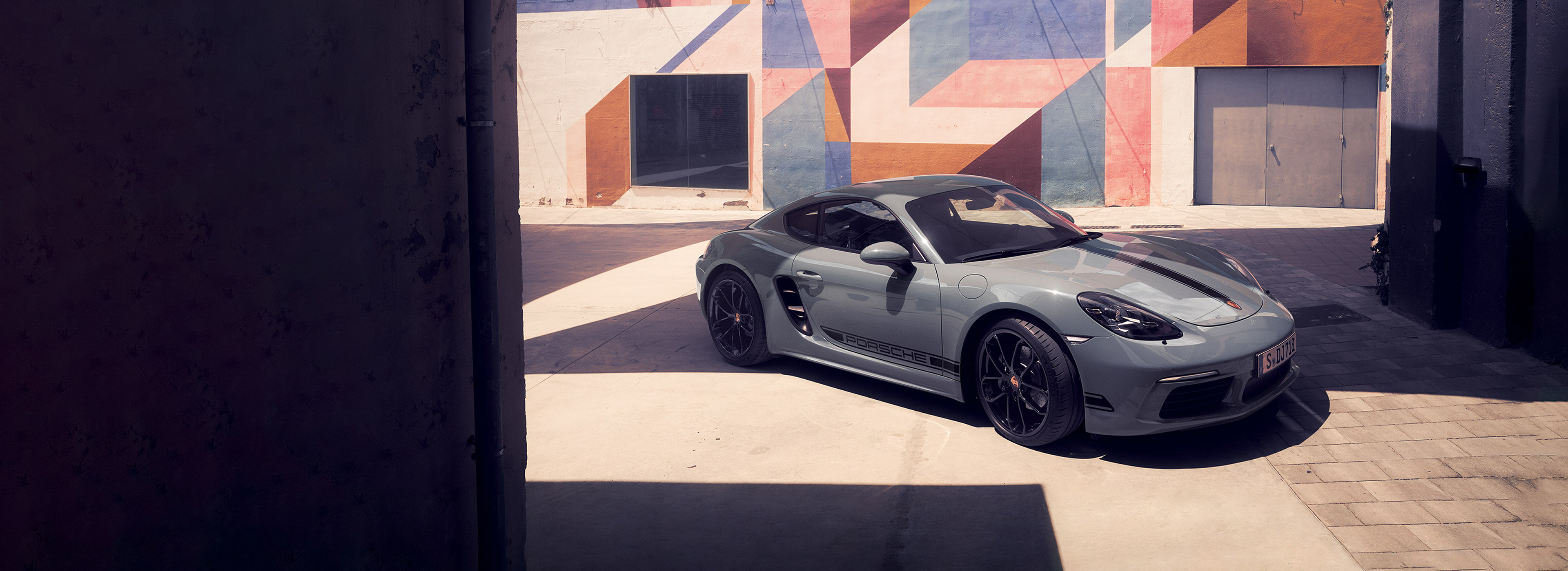 Porsche 718 Cayman parked in a sunlit urban courtyard with geometric mural walls in the background.