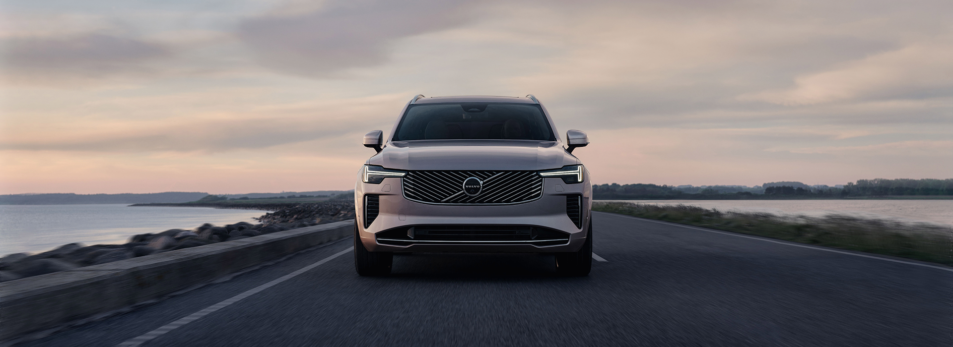 Front view of a gray Volvo SUV driving on a coastal road at sunset, with a serene water body on the side.